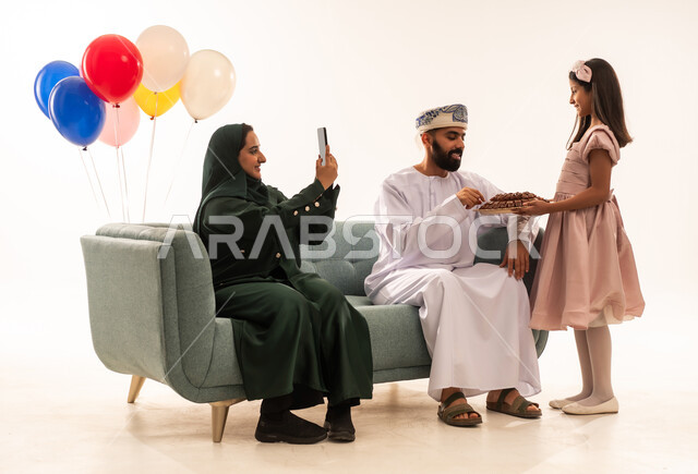 التقاط الصور الشخصية فى العيد , الاحتفال بالأعياد الإسلامية والزيارات العائلية، تقديم الحلويات الشعبية،أجواء البهجة والسعادة في العيد، بورتريه لعائلة عربية خليجية عمانية تجلس في غرفة المعيشة وتقوم بتبادل المعايدات، مفهوم الحنان والمحبة والمودة، خلفية بيضاء .