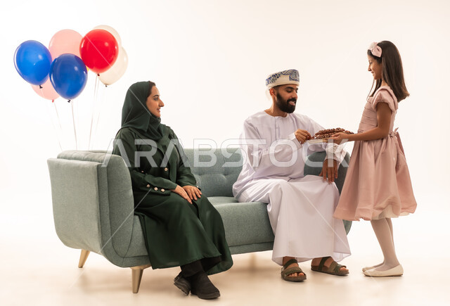 أجواء البهجة والسعادة في العيد، التهنئة والتحية بين أفراد الأسرة, الاحتفال بالأعياد الإسلامية والزيارات العائلية، بنت عربية خليجية عمانية ترتدي ثياب العيد تقوم بتقديم الحلويات الشعبية، بورتريه لعائلة عربية خليجية عمانية تجلس في غرفة المعيشة وتقوم بتبادل المعايدات، مفهوم الحنان والمحبة والمودة، خلفية بيضاء .