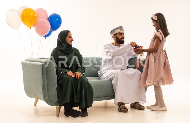 Congratulations and greetings between family members, an atmosphere of joy and happiness on Eid, celebrating Islamic holidays and family visits, an Arab Gulf Omani girl wearing Eid clothes offering traditional sweets, a portrait of an Arab Gulf Omani family sitting in the living room exchanging greetings, the concept of tenderness, love and affection, white background.