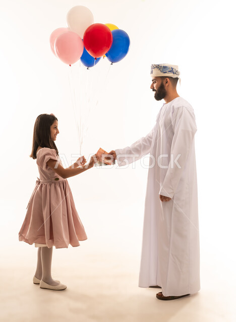 تقديم العيدية للأطفال في عيد الفطر المبارك ، بورتريه لاب عربي خليجي عماني يحتفل بعيد الفطر السعيد مع ابنته،  تقديم العيديات و الحلوى ، جو البهجة والفرح والزينة ، التهنئة والتحية بين أفراد الأسرة, عيد فطر مبارك ، خلفية بيضاء .