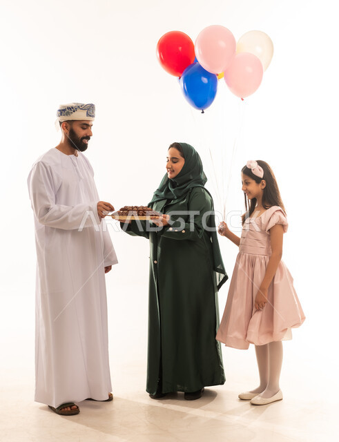 توزيع الحلويات الشعبية في الأعياد، جو البهجة والفرح والزينة، طقوس الاحتفالات والمناسبات السعيدة في سلطنة عمان، بورتريه لعائلة عربية خليجية عمانية تحتفل بالعيد المبارك، ارتداء الأزياء العمانية التقليدية، مفهوم الكرم وتقديم الضيافة العربية، خلفية بيضاء .