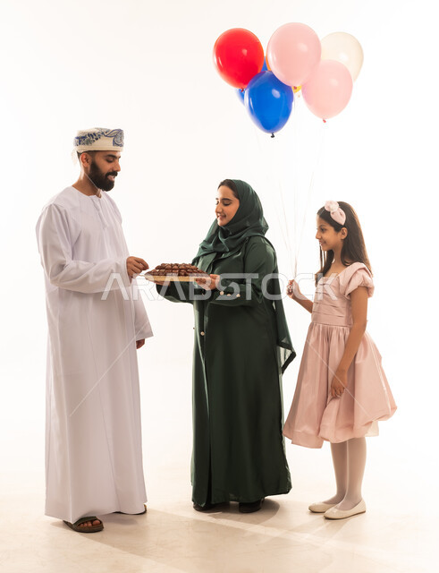 جو البهجة والفرح والزينة، طقوس الاحتفالات والمناسبات السعيدة في سلطنة عمان، توزيع الحلويات الشعبية في الأعياد، بورتريه لعائلة عربية خليجية عمانية تحتفل بالعيد المبارك، ارتداء الأزياء العمانية التقليدية، مفهوم الكرم وتقديم الضيافة العربية، خلفية بيضاء .
