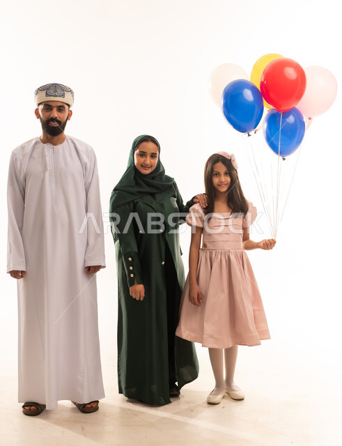 البلالين الملونة والزينة، السلام والتهنئة بقدوم العيد, إظهار مشاعر المودة والمحبة العائلية, أجواء البهجة والسعادة والاحتفال, بورتريه لعائلة عربية خليجية عمانية تحتفل بالأعياد الاسلامية, تقديم الحلويات التقليدية والمأكولات الشهية, خلفية بيضاء .