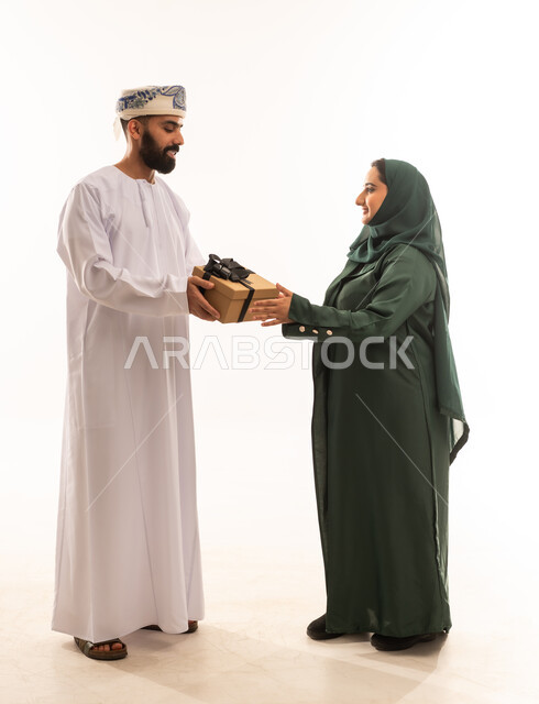 تقديم الهدايا في العيد والمناسبة الخاصة، التعبير عن الحب والتقدير والاحترام، بورتريه لرجل عربي خليجي عماني يرتدي الدشداشة والكمة يقدم صندوق الهدية إلى زوجته بإيماءات السرور والسعادة، مفهوم الشكر والامتنان، الجسم بالطول الكامل، خلفية بيضاء .