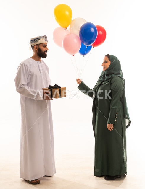التعبير عن الحب والتقدير والاحترام، تقديم الهدايا في العيد والمناسبة الخاصة، بورتريه لرجل عربي خليجي عماني يرتدي الدشداشة والكمة يقدم صندوق الهدية إلى زوجته بإيماءات السرور والسعادة، مفهوم الشكر والامتنان، الجسم بالطول الكامل، خلفية بيضاء .