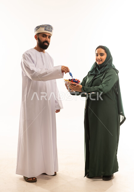 كرم الضيافة وحسن الاستقبال ، طقوس الاحتفالات والمناسبات السعيدة في سلطنة عمان، جو البهجة والفرح والزينة، بورتريه لزوجان عربيان خليجيان عمانيان يحتفلان بالعيد المبارك، ارتداء الأزياء العمانية التقليدية، توزيع الحلويات الشعبية في الأعياد، خلفية بيضاء .