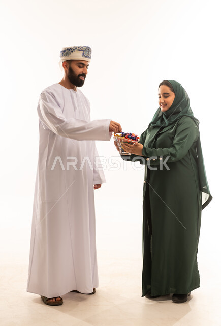 An atmosphere of joy, happiness, and decorations. A portrait of an Omani Gulf Arab couple celebrating Eid al-Adha. Generous hospitality and warm welcome. The rituals of celebrations and happy occasions in the Sultanate of Oman. Wearing traditional Omani costumes. Distributing traditional sweets during the holidays. White background.