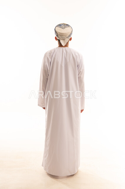 الاهتمام بالأناقة والمظهر الخارجي, مفهوم الرجولة والثقة بالنفس، بورتريه من الخلف لرجل عربي خليجي عماني يرتدي الدشداشة و الكمه ينظر الى الخلف بإيماءات السرور والسعادة، صورة الجسم بالطول الكامل, خلفية بيضاء .