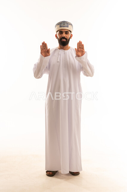 The concept of refusal, abstention and annoyance, a portrait of an Arab Gulf Omani man wearing a dishdasha and a kuma, extending his hand forward with his palms raised in a gesture of forbidding something, asking to stop and not approach, expressions of firmness and seriousness, a full-length body image, a white background.
