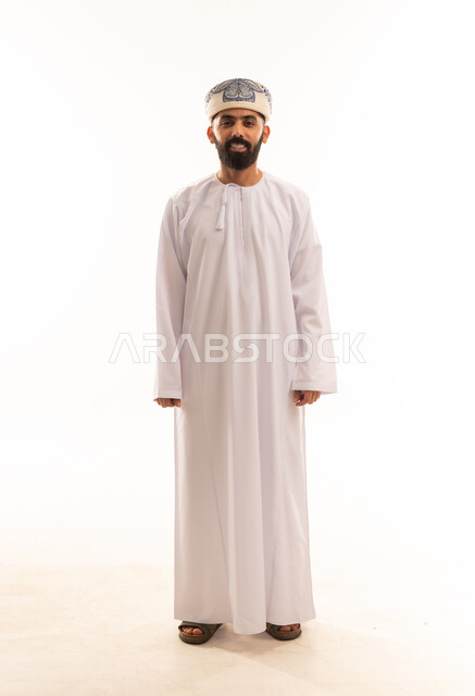 التقاط الصور الشخصية للمناسبات العامه، الاهتمام بالأناقة والمظهر الخارجي, مفهوم الرجولة والثقة بالنفس، بورتريه لرجل عربي خليجي عماني مبتسم يرتدي الدشداشة و الكمه ينظر الى الكاميرا بإيماءات السرور والسعادة، صورة الجسم بالطول الكامل, خلفية بيضاء .