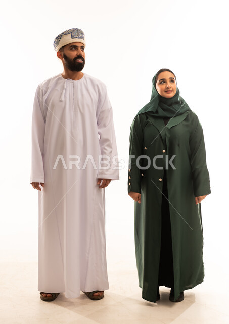 تحديد هدف معين فى الاعلى ، اجواء مليئة بالحب ، قضاء اوقات عائلية ممتعة ومسلية، بورتريه لرجل عربي خليجي عماني يرتدي الدشداشة والكمة يقف مع زوجته بايماءات السعادة والسرور، تعزيز العلاقات الاجتماعية، الجسم بالطول الكامل، خلفية بيضاء .