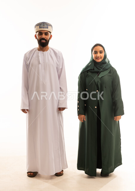 قضاء اوقات عائلية ممتعة ومسلية، اجواء مليئة بالحب ، بورتريه لرجل عربي خليجي عماني يرتدي الدشداشة والكمة يقف مع زوجته بايماءات السعادة والسرور، تعزيز العلاقات الاجتماعية، الجسم بالطول الكامل، خلفية بيضاء .