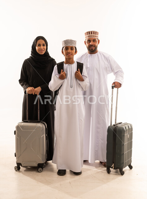 مفهوم السياحة والسفر، الإستعداد لقضاء الإجازة الصيفية والذهاب في رحلة سفر ممتعة، بورتريه لرجل عربي خليجي عماني يرتدي الدشداشة والكمة يقف مع زوجته وابنه ويمسك حقيبة السفر، الأجواء العائلية المليئة بالحب، الجسم بالطول الكامل، خلفية بيضاء .