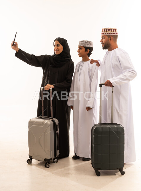 توثيق اللحظات التذكارية باستخدام الجوال ، الأجواء العائلية المليئة بالحب، بورتريه لرجل عربي خليجي عماني يرتدي الدشداشة والكمة يقف مع زوجته وابنه ويمسك حقيبة السفر،تنظر الى الاعلى بايماءات السعادة ،الإستعداد لقضاء الإجازة الصيفية والذهاب في رحلة سفر ممتعة، الجسم بالطول الكامل، خلفية بيضاء .
