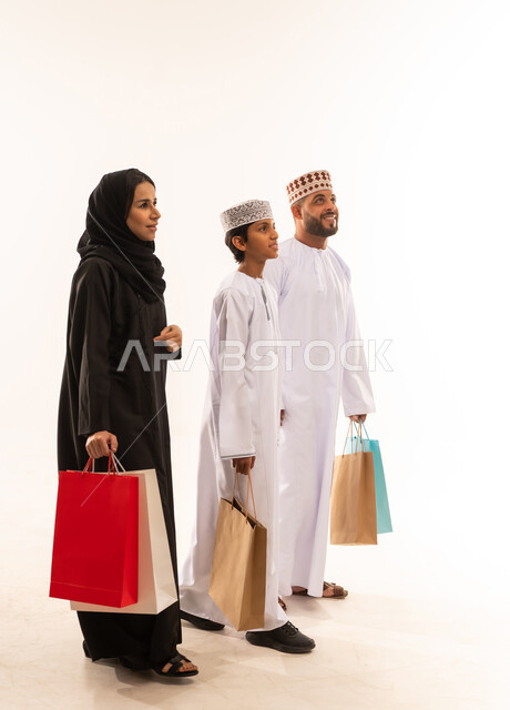 الاستفادة من الخصومات على المنتجات، ايماءات السرور بعد التسوق والتبضع مع العائلة ، تخفيضات وعروض موسمية في الأسواق العمانية,  بورتريه من الجانب لعائلة عربية خليجية عمانية يرتدي الملابس العمانية التقليدية تحمل أكياس تسوق ملونه ، صورة الجسم بالطول الكامل، خلفية بيضاء .