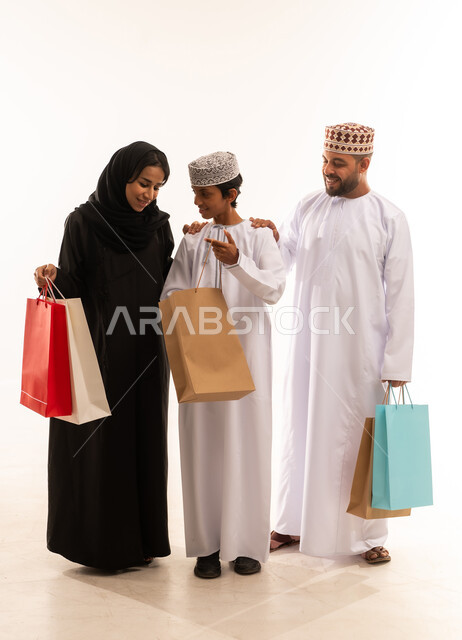 شراء الاغراض الضرورية ، ايماءات السرور بعد التبضع مع العائلة ، تخفيضات وعروض موسمية في الأسواق العمانية, بورتريه لعائلة عربية خليجية عمانية يرتدي الملابس العمانية التقليدية تحمل أكياس تسوق ملونه ، صورة الجسم بالطول الكامل، خلفية بيضاء .