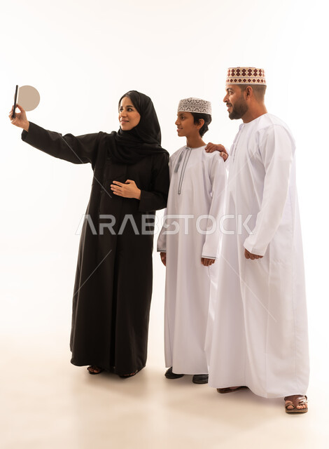 مفهوم الأمان والإستقرار العائلي، إجراء مكالمة فيديو أونلاين، توثيق اللحظات العائلية السعيدة، ايماءات القاء التحية والسلام، بورتريه لعائلة عربية خليجية عمانية تلتقط صورة تذكارية من خلال الهاتف المحمول، المحافظة على الترابط الأسري، خلفية بيضاء .