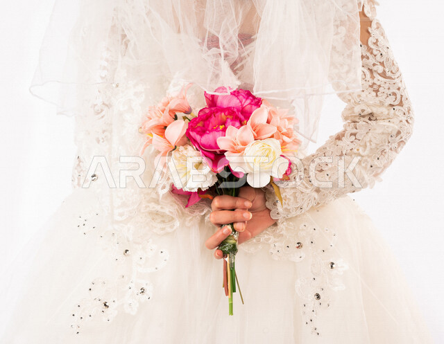 Angled portrait from the back of a Saudi Arabian Gulf bride, a wonderful photo session of a Saudi Arab bride on her wedding day, wearing a white wedding suit, holding a beautiful bouquet of roses in her hand, the night of life, occasions and weddings