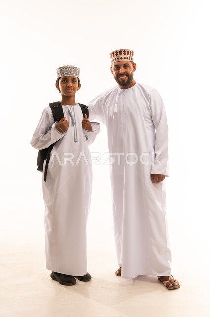 Reaching the highest ranks through hard work and perseverance, preparing to start a new school year, the concept of learning in advanced schools in the Sultanate of Oman, a portrait of an Arab Gulf Omani father wearing a dishdasha and a kummah helping his son get ready to go to school, full body, white background.