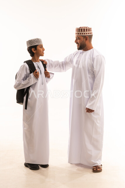 الاستعداد لبدء عام دراسي جديد، الوصول الى المراتب العليا بالاجتهاد والمثابرة، مفهوم التعلم في المدارس المتطورة في سلطنة عمان، بورتريه لأب عربي خليجي عماني ترتدي الدشداشة والكمة يقوم بمساعدة ابنه بالتجهز للذهاب الى المدرسة، الجسم بالطول الكامل، خلفية بيضاء .