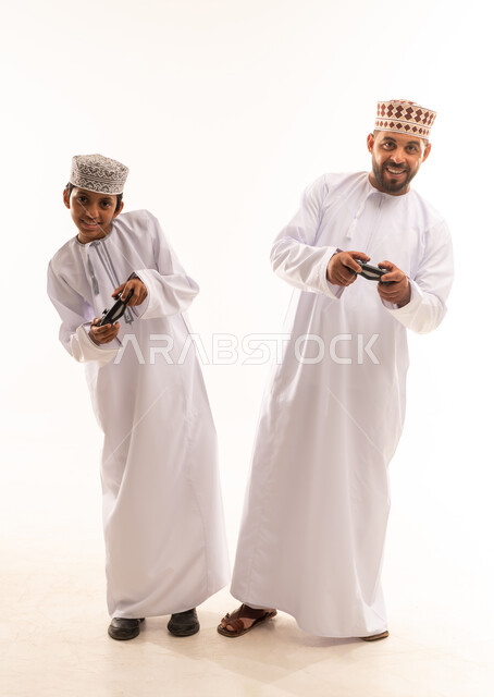 التفاعل والاندماج مع ألعاب الفيديو، قضاء وقت ممتع ومسلي، اجهزة وتقنيات الكترونية ترفيهيه حديثة, بورتريه لولد عربي خليجي عماني يرتدي الدشداشة والكمة يقف مع ابيه ويمسك عصا التحكم، صورة لكامل الجسم، خلفية بيضاء .