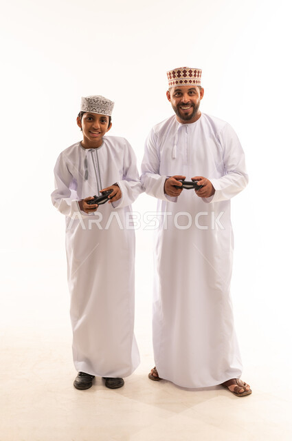 قضاء وقت ممتع ومسلي، اجهزة وتقنيات الكترونية ترفيهيه حديثة, التفاعل والاندماج مع ألعاب الفيديو، بورتريه لولد عربي خليجي عماني يرتدي الدشداشة والكمة يقف مع ابيه ويمسك عصا التحكم، صورة لكامل الجسم، خلفية بيضاء .