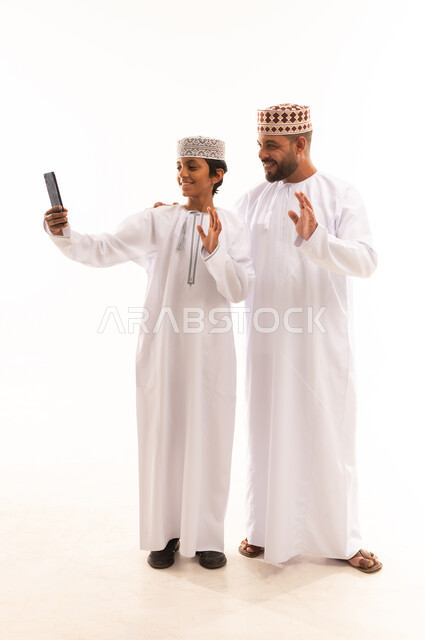 ايماءات القاء التحية والسلام، اجراء مكالمة فيديو أونلاين، توثيق اللحظات العائلية السعيدة، استخدام جهاز تقني حديث ومتطور, بورتريه لولد عربي خليجي عماني يرتدي الدشداشة والكمة يقف مع أبيه ويمسك بيده الجوال, الجسم بالطول الكامل, خلفية بيضاء .