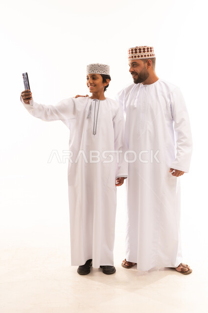 Making an online video call, documenting happy family moments, greeting and peace gestures, using a modern and advanced technical device, portrait of an Arab Gulf Omani boy wearing a dishdasha and a kummah, standing with his father and holding a mobile phone, full body, white background.