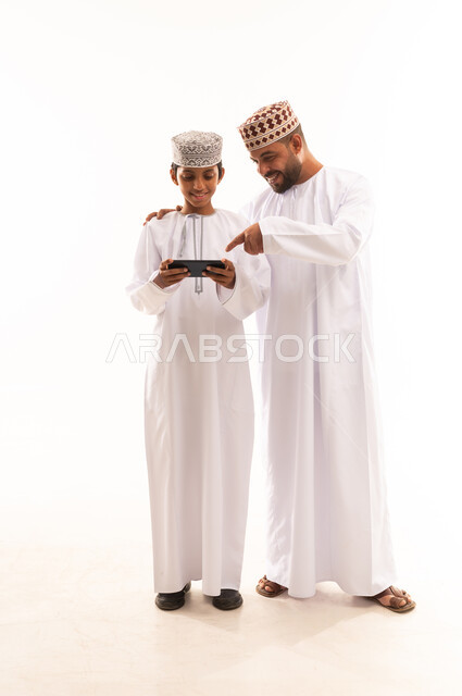 مشاهدة الأفلام والمسلسلات في أوقات الفراغ، استخدام جهاز تقني حديث ومتطور, بورتريه لولد عربي خليجي عماني يرتدي الدشداشة والكمة يقف مع أبيه ويمسك بيده الجوال, الجسم بالطول الكامل, خلفية بيضاء .