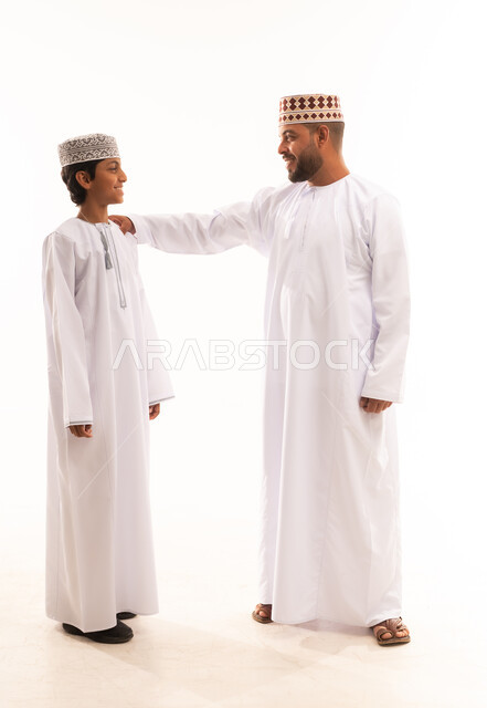 أجواء أسرية مليئة بالمودة، الرقي في التعامل وتبادل الأحاديث، بورتريه لأب وابنه عربيان خليجيان عمانيان يرتديان الدشداشة والكمة ، ارتداء الملابس العمانية التقليدية في الأعياد الإسلامية، خلفية بيضاء .