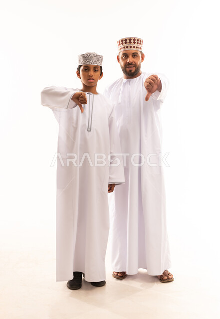 حركة يد وايماءات وجه تدل على عدم الإعجاب والموافقة، الشعور بالإنزعاج من شيء ما، الرابطة الأبوية والترابط الأسري، بورتريه لأب وابنه عربيان خليجيان عمانيان يرتديان الدشداشة والكمة ، ارتداء الملابس العمانية التقليدية في الأعياد الإسلامية، خلفية بيضاء .