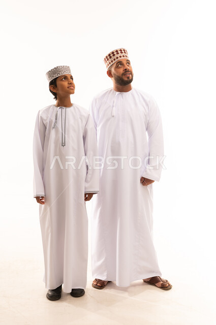 النظر الى شئ ما بايماءات السعادة ، حب الأب الكبير لأولاده , الرابطة الأبوية والترابط الأسري، ايماءات الحب والعطف على الأبناء، بورتريه لأب وابنه عربيان خليجيان عمانيان يرتديان الدشداشة والكمة ، ارتداء الملابس العمانية التقليدية في الأعياد الإسلامية، خلفية بيضاء .