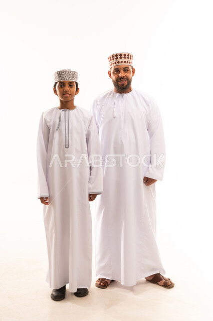 ارتداء الملابس العمانية التقليدية في الأعياد الإسلامية، النظر الى الكاميرا بايماءات السعادة ، حب الأب الكبير لأولاده , الرابطة الأبوية والترابط الأسري، ايماءات الحب والعطف على الأبناء، بورتريه لأب وابنه عربيان خليجيان عمانيان يرتديان الدشداشة والكمة ، خلفية بيضاء .