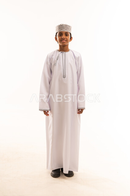 Standing up straight and confident, the concept of elegance and interest in one's outward appearance, a portrait of a smiling Omani Gulf Arab boy wearing a dishdasha and a kummah, looking at the camera with an expression of happiness and pleasure, white background.