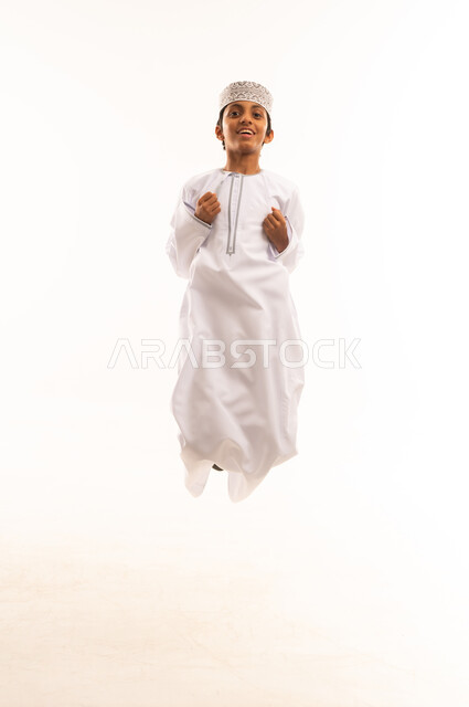 مفهوم اللهو واللعب, بورتريه لولد عربي خليجي عماني يرتدي الدشداشة و الكمه يتظهر عليه علامات السعادة، القفز في الجو تعبيرا عن الفرح والسرور, صورة الجسم بالطول الكامل, خلفية بيضاء .
