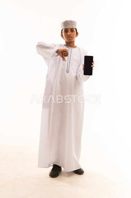 استخدام جهاز تقني حديث ومتطور، عرض شاشة سوداء فارغة، الدردشة والتواصل مع الاهل والاصدقاء عبر الجوال، بورتريه لولد عربي خليجي عماني يرتدي الدشداشه والكمه يمسك الهاتف المحمول ينظر إلى الكاميرا بإيماءات الرفض ، الجسم بالطول الكامل، خلفيه بيضاء .