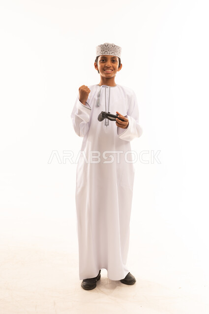 Using technological devices and modern techniques, gestures of enjoyment and integration, spending fun times playing video games, portrait of an Arab Gulf Omani boy wearing a dishdasha and a kuma holding a joystick, white background