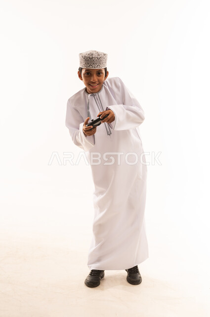 إيماءات الاستمتاع والاندماج، قضاء أوقات ممتعة في ألعاب الفيديو الترفيهيه، بورتريه لولد عربي خليجي عماني يرتدي الدشداشة والكمة يحمل بيده عصا التحكم، استخدام الأجهزة التكنولوجية والتقنيات الحديثة، خلفية بيضاء