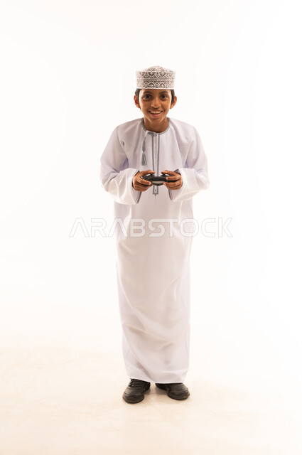 قضاء أوقات ممتعة في ألعاب الفيديو الترفيهيه، بورتريه لولد عربي خليجي عماني يرتدي الدشداشة والكمة يحمل بيده عصا التحكم، إيماءات الاستمتاع والاندماج، استخدام الأجهزة التكنولوجية والتقنيات الحديثة، خلفية بيضاء
