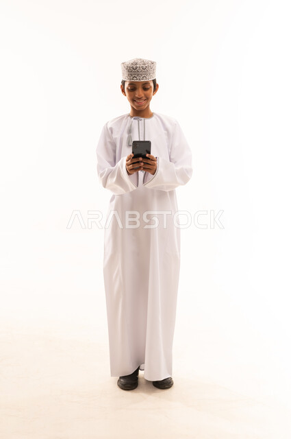 تبادل الأخبار مع الأهل والأصدقاء، إجراء مكالمة هاتفية باستخدام الموبايل،  بورتريه لولد عربي خليجي عماني يرتدي الدشداشة والكمه يقوم بالتواصل مع الأقارب، تعابير الفرح والسرور، صورة الجسم بالطول الكامل, خلفية بيضاء
