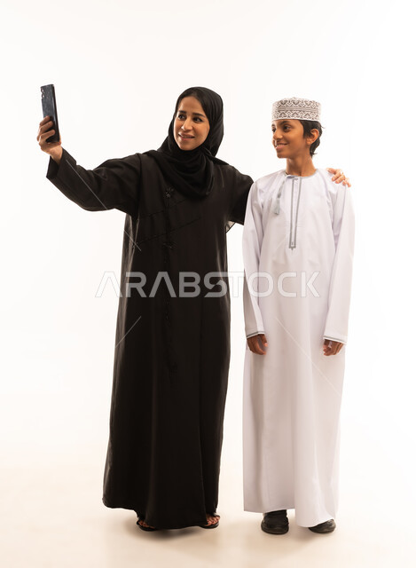 توثيق اللحظات المميزة باستخدام الأجهزة التقنية الحديثة, فعاليات و نشاطات مع الأسرة, قضاء الأوقات الممتعة مع الأبناء, بورترية لامرأة عربية خليجية عمانية مع ابنها يرتديان الزي التقليدي العماني يقومان بالتقاط صور السيلفي التذكارية عبر الجوال, الأجواء العائلية السعيدة .