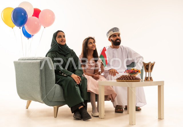 Omani family gathering to welcome Eid, showing feelings of joy and happiness at the arrival of Eid, portrait of an Omani Gulf Arab family sitting in the living room looking at something with happy gestures, exchanging greetings, the concept of tenderness, love and affection, offering Arab hospitality and traditional sweets, white background.