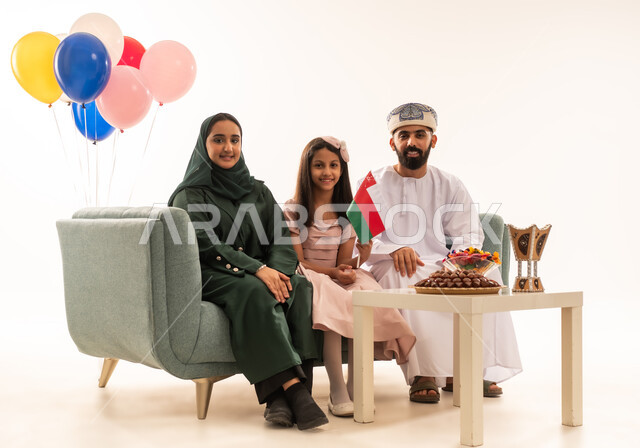 Expressing feelings of joy and happiness with the arrival of Eid, family visits and celebration of Islamic holidays, a portrait of an Arab Gulf Omani family sitting in the living room looking at the camera with happy gestures, exchanging greetings, the concept of tenderness, love and affection, offering Arab hospitality and traditional sweets, white background.