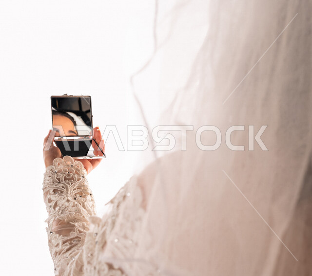 بورتريه بصورة مقربة لإنعكاس صورة عروسة عربية خليجية سعودية جميلة في مرآة صغيرة ، أجمل اطلالة لعروسة عربية سعودية خليجية في يوم زفافها ، ترتدي بدلة الزفاف البيضاء ، ليلة العمر ، مناسبات وأفراح ، خلفية بيضاء