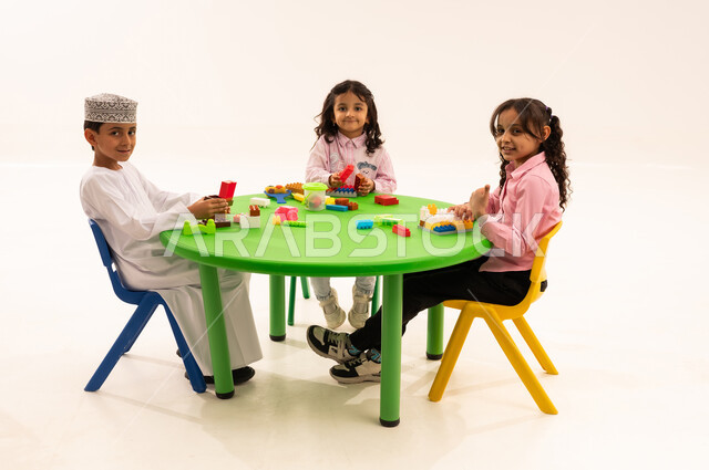 Enhancing mental and intellectual abilities, performing skills and fun activities, developing artistic skills through shaping, educational sessions and classes in Omani kindergartens, a portrait of an Arab Gulf Omani student wearing a kummah and a dishdasha playing with colored cubes with his friends, white background