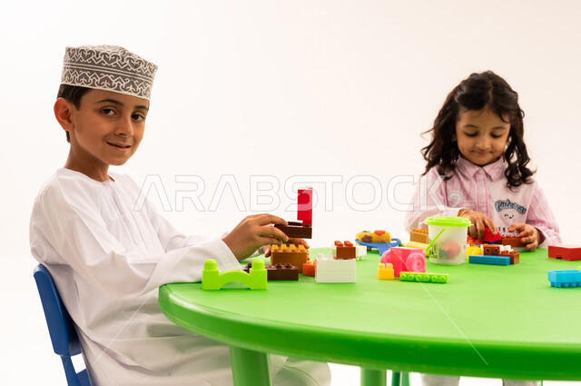 Strengthening mental and intellectual abilities, performing skills and fun activities, developing artistic skills through shaping, educational sessions and classes in Omani kindergartens, a portrait of an Arab Gulf Omani student wearing a kummah and a dishdasha playing with colored cubes with his friends, white background