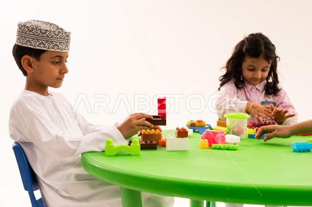 تعزيز القدرات العقلية والذهنية، القيام بالمهارات و الأنشطة المسلية الممتعة، تنيمة المهارات الفنية بالتشكيل، الجلسات والحصص التعليمية في الروضات العمانية، بورتريه لطالب عربي خليجي عماني يرتدي الكمة والدشداشة يقوم باللعب في المكعبات الملونة برفقة اصدقاءه، خلفية بيضاء