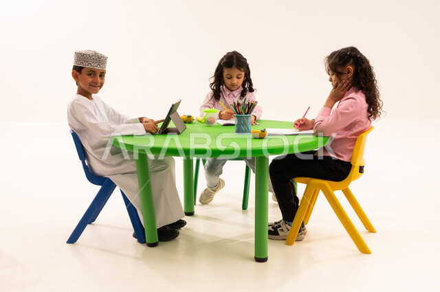 نشاط ترفيهي مسلي، تعزيز المواهب الابداعية الفنية عند الطلاب، تعلم مهارات التركيز والابتكار عبر جهاز التابلت، بورتريه لطالب عربي خليجي عماني يرتدي الدشداشة والكمة يقوم باستخدام جهاز لوحي بجانب اصدقاءه، الرسم والتلوين واستخدام الاجهزة التقنية الحديثة، خلفية بيضاء