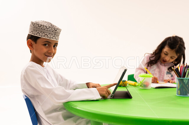 نشاط ترفيهي مسلي، تعزيز المواهب الابداعية الفنية عند الطلاب، تعلم مهارات التركيز والابتكار عبر جهاز التابلت، بورتريه لطالب عربي خليجي عماني يرتدي الدشداشة والكمة يقوم باستخدام جهاز لوحي بجانب اصدقاءه، الرسم والتلوين واستخدام الاجهزة التقنية الحديثة، خلفية بيضاء