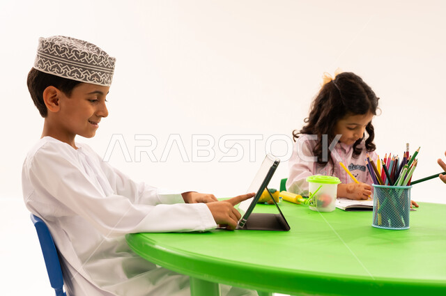 An entertaining and fun activity, enhancing students' artistic creativity, learning concentration and innovation skills through a tablet. A portrait of an Omani Gulf Arab student wearing a dishdasha and a kuma using a tablet with his friends, drawing, coloring, and using modern technical devices, white background.